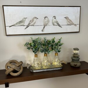 May include: A brown wooden shelf with a painting of five birds on a wire, three clear glass vases with lights inside, a wooden knot, and a stack of gray stones with the words "This is our happy place" on them.