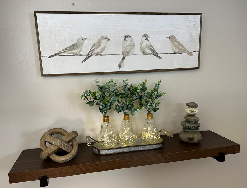 May include: A brown wooden shelf with a painting of five birds on a wire, three clear glass vases with lights inside, a wooden knot, and a stack of gray stones with the words "This is our happy place" on them.