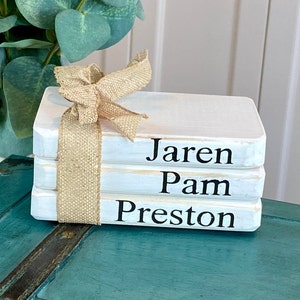 May include: A stack of three white wooden blocks, resembling books, tied with a burlap ribbon. The names "Jaren", "Pam", and "Preston" are printed in black on the sides of the blocks. The blocks are on a teal surface.