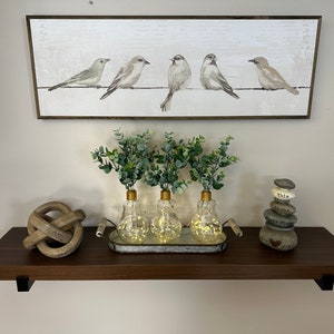 May include: A dark brown wooden shelf with three clear glass light bulb vases with greenery and gold tops. The vases are on a metal tray with a wooden handle. There is a wooden knot sculpture on the left and a stack of gray stones with white lettering on the right. Above the shelf is a framed print of five gray birds on a wire.