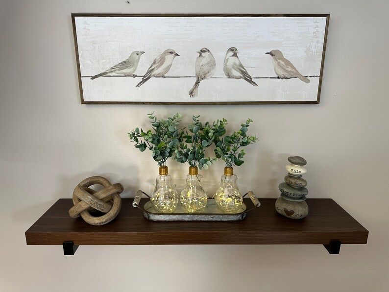 May include: A dark brown wooden shelf with three clear glass light bulb vases with greenery and gold tops. The vases are on a metal tray with a wooden handle. There is a wooden knot sculpture on the left and a stack of gray stones with white lettering on the right. Above the shelf is a framed print of five gray birds on a wire.
