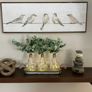 May include: Three clear glass light bulb vases with gold tops and warm white fairy lights inside, filled with green eucalyptus sprigs, on a silver tray with a wooden handle, on a dark brown shelf with a wooden knot and a stack of gray stones with the text "This is our Happy Place" on the shelf.