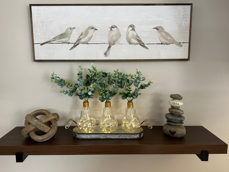 May include: Three clear glass light bulb vases with gold tops and warm white fairy lights inside, filled with green eucalyptus sprigs, on a silver tray with a wooden handle, on a dark brown shelf with a wooden knot and a stack of gray stones with the text "This is our Happy Place" on the shelf.