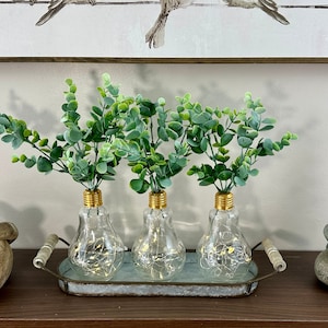 May include: Three clear glass light bulb vases with gold tops, filled with green eucalyptus sprigs, are arranged on a silver tray with handles. The tray is on a wooden surface with a wooden knot and a stack of stones with the text "This is our Happy Place" on a white background.
