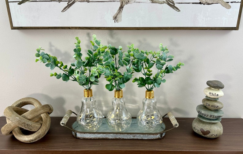 May include: Three clear glass light bulb vases with gold tops, filled with green eucalyptus sprigs, are arranged on a silver tray with handles. The tray is on a wooden surface with a wooden knot and a stack of stones with the text "This is our Happy Place" on a white background.