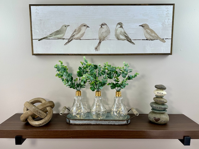 May include: Three clear glass lightbulb vases with gold tops, filled with green eucalyptus sprigs, are arranged on a metal tray with wooden handles. The tray is on a brown shelf with a wooden knot sculpture and a stack of gray stones with the text "This is our Happy Place" on a white background.