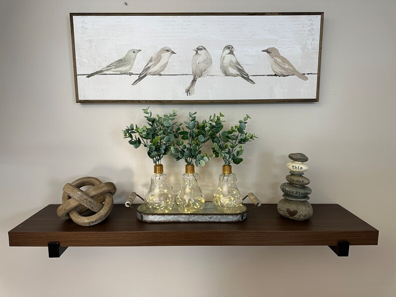 May include: A brown wooden shelf with three glass vases with lights inside. The vases are filled with green eucalyptus branches. The shelf is decorated with a wooden knot and a stack of stones with the words "This is our Happy Place" written on them. A framed painting of five birds on a wire is hanging on the wall above the shelf.