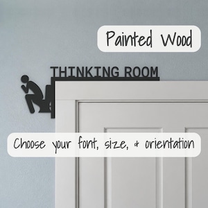 May include: A black wooden door corner sign featuring the text THINKING ROOM and a silhouette of a person sitting on a toilet. The sign is mounted on the upper corner of a white door frame, displaying a flat, matte finish design.