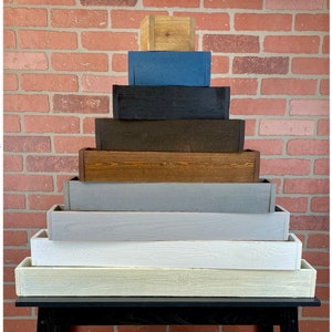 May include: A set of nine wooden planter boxes stacked in a pyramid shape. The boxes are painted in various colors, including natural wood, brown, black, blue, gray, and white. The boxes are labeled with the number of jars they can hold and their width in inches. The boxes are displayed on a black table against a brick wall.