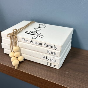 May include: A stack of four white wooden books with black text. The top book says "Love" in cursive. Below, the books read "The Wilson Family", "Kirk", "Alysha", and "Ellie". A jute twine and wooden bead accent is tied around the books.