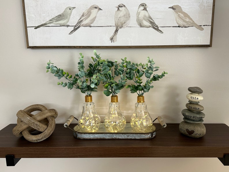 May include: Three clear glass light bulb vases with gold tops, filled with green eucalyptus sprigs, are arranged on a metal tray. The tray is on a dark brown shelf with a wooden knot and a stack of stones with the words "This is our Happy Place" on a white background.