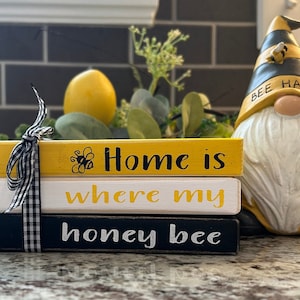 May include: Three wooden blocks stacked on top of each other. The top block is yellow and says "Home is" with a small bee illustration. The middle block is white and says "where my" in yellow lettering. The bottom block is black and says "honey bee" in white lettering. The blocks are tied together with a black and white checkered ribbon.