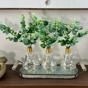 May include: Three clear glass vases with gold tops, filled with green leafy stems, are arranged on a rustic metal tray. The vases are shaped like light bulbs and have a warm glow emanating from inside.