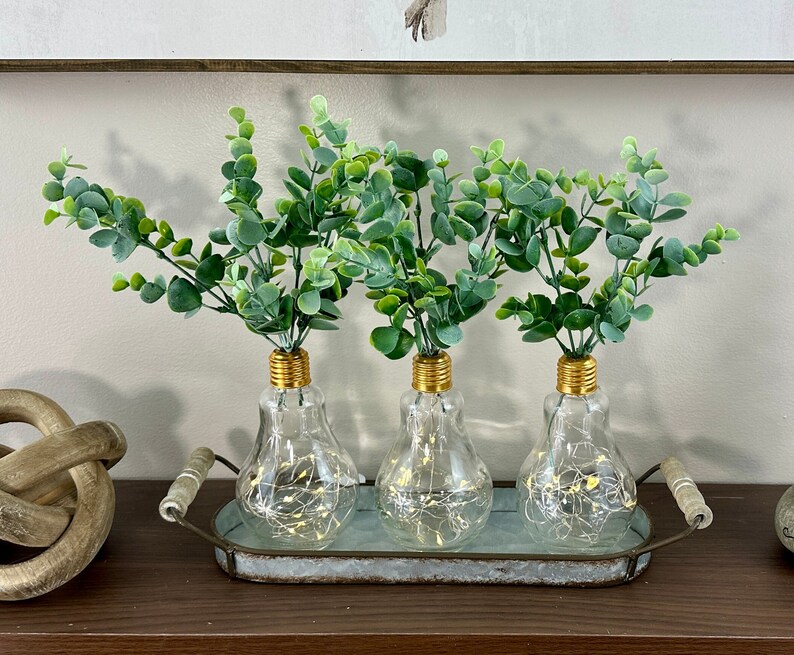 May include: Three clear glass vases with gold tops, filled with green leafy stems, are arranged on a rustic metal tray. The vases are shaped like light bulbs and have a warm glow emanating from inside.