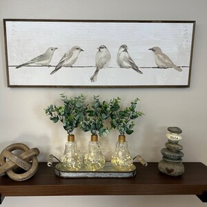 May include: A wooden shelf with a framed print of five birds on a wire, three glass light bulb vases with greenery, a wooden knot, and a stack of painted stones with words on them.