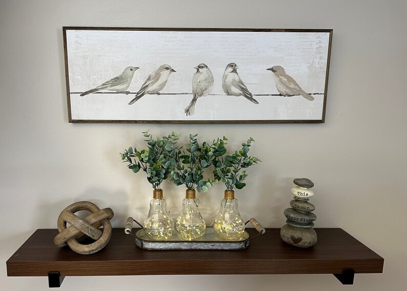 May include: A wooden shelf with a framed print of five birds on a wire, three glass light bulb vases with greenery, a wooden knot, and a stack of painted stones with words on them.