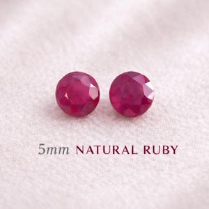 Könnte beinhalten: Zwei runde, facettierte, natürliche Rubine, jeweils 5 mm Durchmesser, auf weißem Stoff. Die Rubine haben eine tiefe, satte rote Farbe. Der Text unten lautet "5mm NATURAL RUBY".