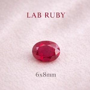 Könnte beinhalten: Ein oval geschliffener Labor-Rubin, 6x8mm groß, in tiefroter Farbe, auf einem weichen, strukturierten Hintergrund. Über dem Edelstein steht der Text "LAB RUBY". Die Facetten des Rubins reflektieren das Licht.