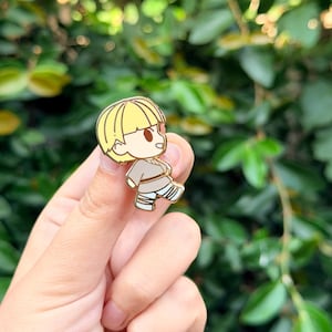 Puede incluir: Un pequeño pin de esmalte de estilo caricatura que representa a un personaje con cabello amarillo y un atuendo beige. El pin se sostiene entre los dedos sobre un fondo de follaje verde. El personaje tiene una expresión amigable.