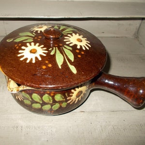 Può includere: Una pentola in ceramica marrone con coperchio, con un motivo floreale in bianco, verde e arancione. La pentola ha un manico e un beccuccio.
