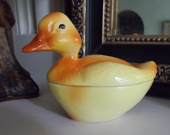 May include: A ceramic duck shaped dish with a yellow and orange glaze. The duck has a closed beak and black eyes.