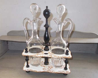 May include: A vintage glass oil and vinegar cruet set with a white and brown ceramic stand. The cruets have black wooden stoppers and are on a stand with black wooden legs.