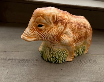 May include: A ceramic figurine of a pig in shades of orange and brown. The pig is lying down on a bed of green grass. The figurine has detailed fur texture and is likely a decorative item.