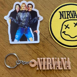 May include: A set of Nirvana merchandise featuring a sticker with a photo of the band, a yellow patch with the band's name and a smiley face logo, and a keychain with the band's name.