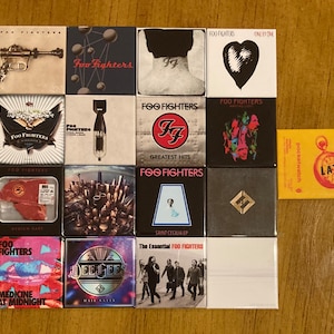 May include: A collection of 12 square magnets featuring various album covers from the band Foo Fighters. The magnets are arranged in a grid of four rows and three columns. The album covers include "One by One", "Greatest Hits", "Saint Cecilia EP", "The Essential Foo Fighters", "Medicine at Midnight", "Hail Satin", "Medium Rare", "Waiting Light", "In Your Honor", "There Is Nothing Left to Lose", "Echoes, Silence, Patience & Grace", and "Concrete and Gold".