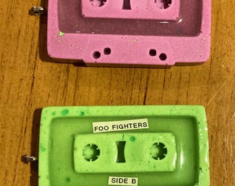 Puede incluir: Dos colgantes de cinta de casete. El superior es rosa con motas verdes y presenta la palabra NIRVANA escrita con cuentas de letras plateadas. El inferior es verde lima con motas verde oscuro, etiquetado con FOO FIGHTERS y el texto SIDE B.