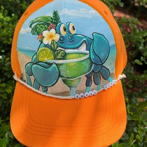 Könnte beinhalten: Orangefarbene Baseballkappe mit einer gemalten Szene eines blauen Krebses, der eine Margarita an einem Strand genießt. Auf der Krempe steht der Text "SHELL YEAH". Der Krebs hält eine Margarita mit Limette und tropischen Blumen.