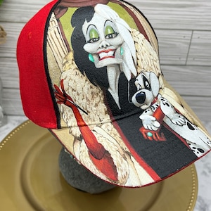 May include: A red baseball cap with a printed image of Cruella De Vil from Disney's 101 Dalmatians. Cruella is wearing a fur coat and holding a cigarette. A Dalmatian puppy is sitting on her shoulder.