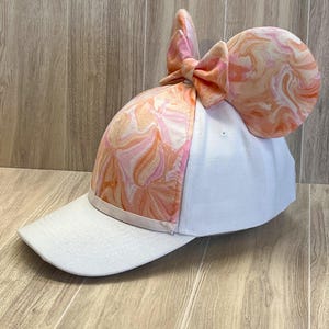 May include: A white baseball cap with a pink and orange marbled fabric bow and ears on top. The fabric has a swirled pattern.