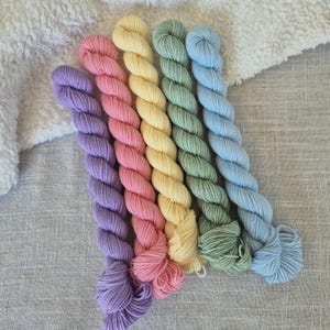 May include: A collection of six skeins of yarn in pastel colors: lavender, pink, yellow, green, and blue. The yarn is arranged in a row, with each skein tightly twisted. The background is a light gray fabric and a white, fluffy material.