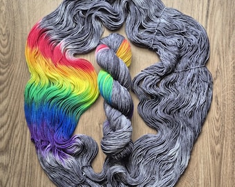 Può includere: Una matassa di filato con un gradiente di colori arcobaleno che include rosso, arancione, giallo, verde, blu e viola, in contrasto con una base di fibre grigie e bianche macchiettate. Il filato è disposto in un grande anello circolare e una matassa.