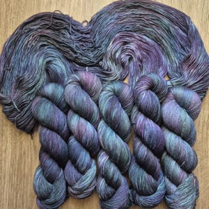 May include: A collection of hand-dyed yarn in shades of blue, purple, and teal. The yarn is arranged in skeins and hanks, showcasing the color variations. The skeins are in a row, and the hanks are in a heart shape.