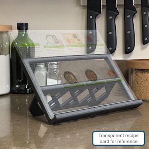 May include: A gray metal recipe stand with a transparent recipe card holder. The stand has a slanted surface and a base with a grid pattern. The recipe card holder is made of clear plastic and has a slot for holding a recipe card. The stand is sitting on a countertop with other kitchen items.