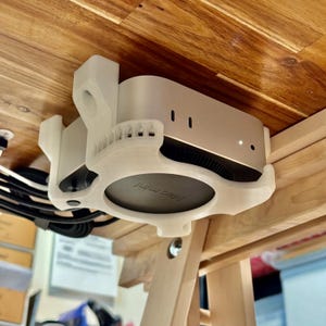 May include: A silver Mac Mini computer is mounted under a wooden table with a white 3D-printed bracket. The bracket securely holds the computer, with the "Mac mini" logo visible. Cables are visible.