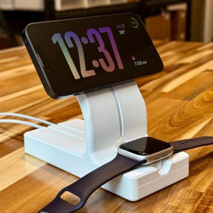 May include: White charging stand with a black and silver Apple Watch and a white iPhone displaying the time 12:37. The iPhone is in a vertical stand and the watch is in a horizontal stand.