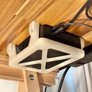 May include: A black electronic device is mounted under a wooden table with a white 3D-printed bracket. The bracket has a geometric design and is secured with screws. Black cables are plugged into the device.