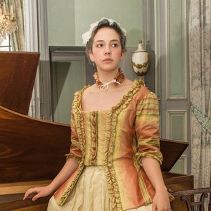 May include: A woman wearing a white lace cap and a striped, ruffled, yellow and orange jacket over a cream-colored dress. She is standing in front of a wooden harpsichord.