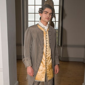 May include: A man wearing a gray coat with gold buttons over a gold vest and white stockings. He is wearing black buckled shoes.