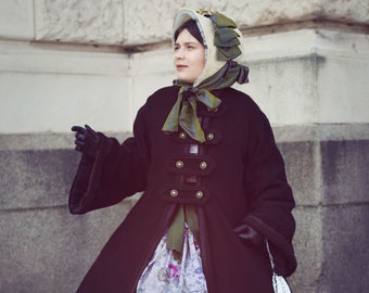 May include: A woman in a vintage black coat with gold buttons and a long, tiered floral skirt. She wears a bonnet with green accents and black gloves, holding a white purse. The outfit is reminiscent of the Victorian era.