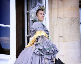 May include: A woman in a gray dress with green trim and a white bonnet stands in front of a white door. She is holding a black umbrella.