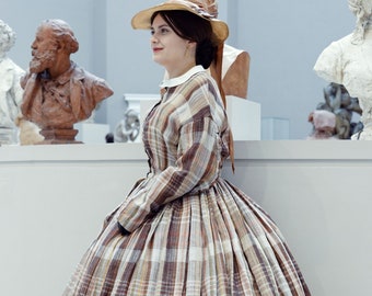May include: A woman wearing a brown and white plaid dress with a ruffled hem and a brown hat. She is standing in a museum setting with sculptures in the background.