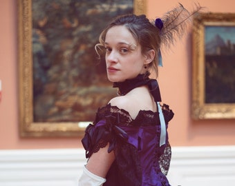 May include: A woman in a black and purple dress with a lace bodice and a feathered hairpiece. She is holding a black fan and is standing in front of a painting.