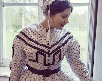 May include: A woman wearing a white Victorian dress with a black floral pattern and a black ribbon around the waist. She is wearing a white lace bonnet and has her hair pulled back. The dress has a black trim around the neckline and sleeves. The woman is standing in front of a window.