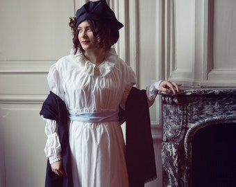 Puede incluir: Una mujer con un vestido blanco, un chal negro y una cinta azul alrededor de la cintura. Lleva un gorro negro y está de pie frente a una chimenea.