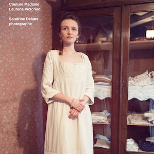 May include: A white, long-sleeved dress with a gathered skirt and a square neckline. The dress is worn by a person standing in front of a wooden cabinet with glass doors. The cabinet is filled with white linens.