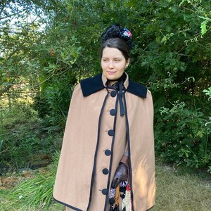 May include: A woman wearing a brown cape with black trim and a black ribbon around her neck. She is holding a black umbrella and a pair of gloves.